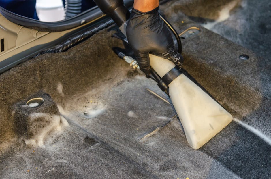 A person vacuuming a large, ornate area rug with floral patterns in shades of beige, green, and orange. The individual, dressed in casual clothing including a beige coat, jeans, and white sneakers, is